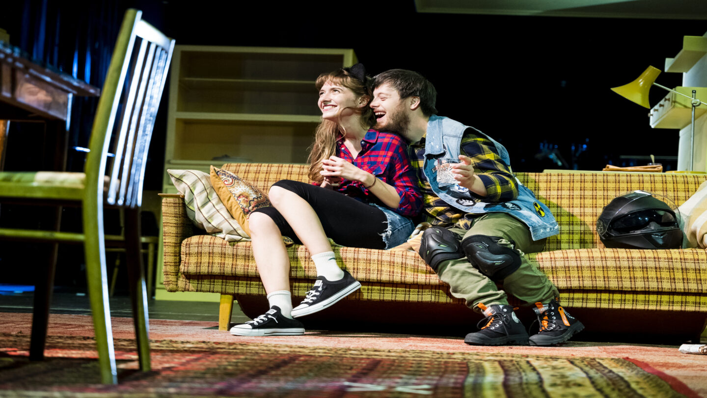 Zdjęcie. Aktorka grająca Siostrę i aktor z zespołem Downa grający Chłopaka siedzą na kanapie i patrzą się w lewą stronę. Chłopak próbuje pocałować aktorkę w szyję.
