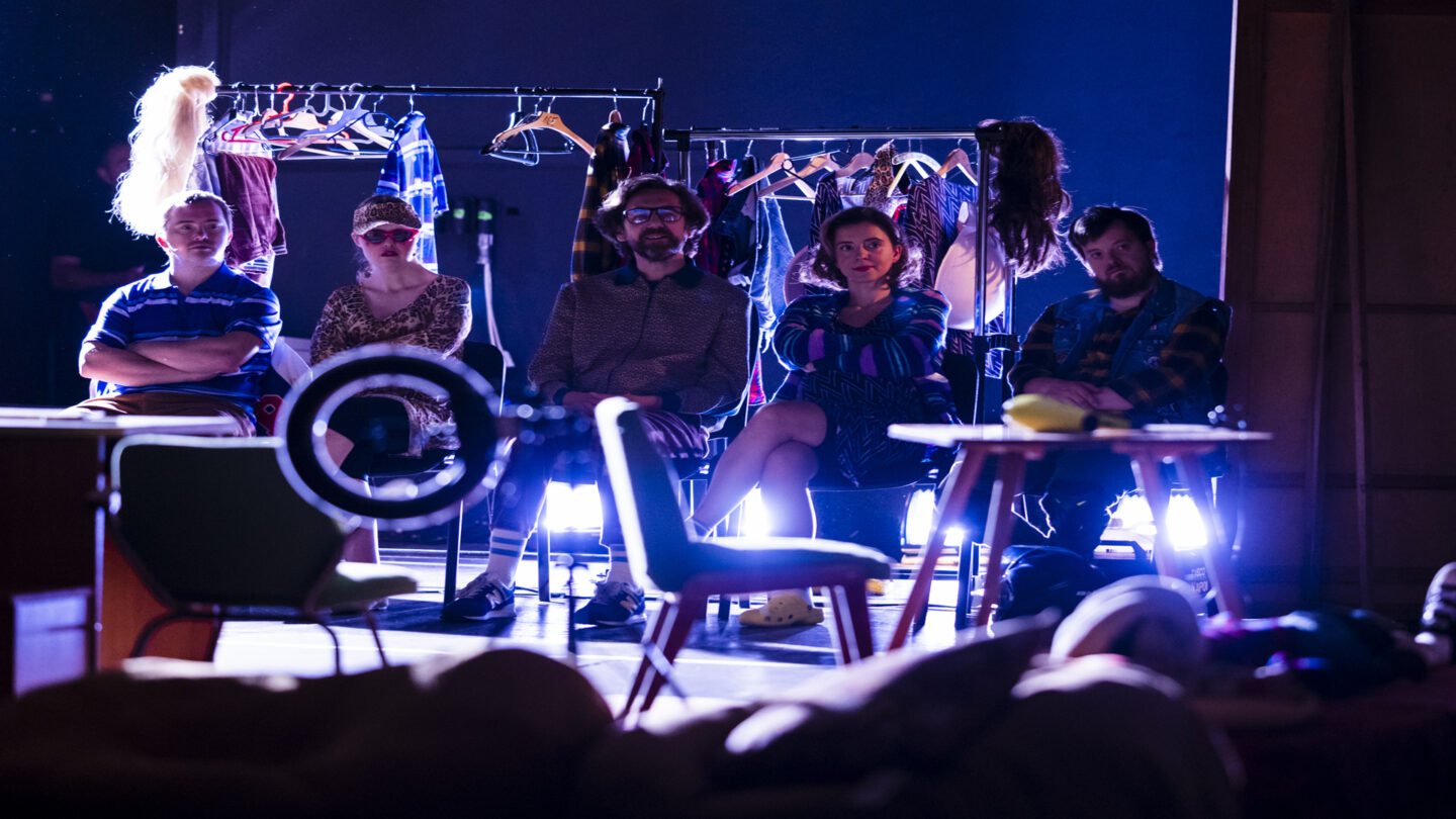 Zdjęcie. Piątka aktorów w kostiumach siedzi na krzesłach, na tyle sceny. Za nimi wieszaki z kostiumami i perukami. Fioletowe światło na podłodze, za nimi.
